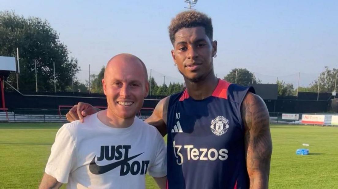 Ryan Hopper and Marcus Rashford pose for a photograph at a football ground with their arms around each other