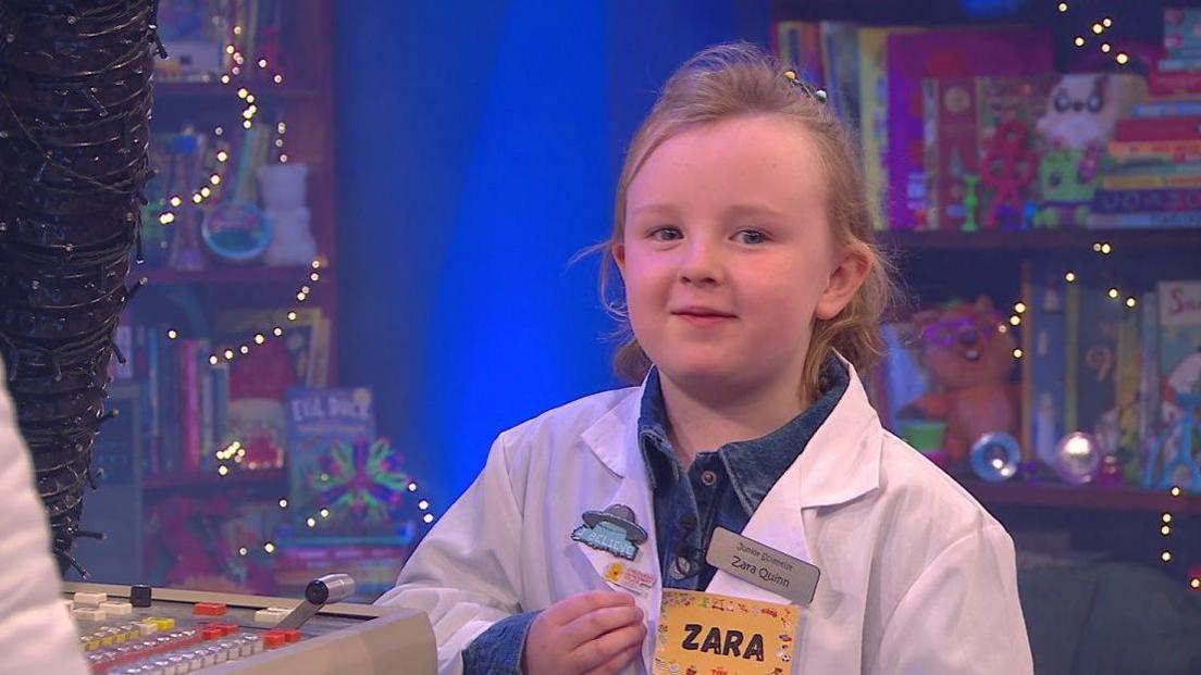 Zara has light brown/blond hair and is wearing a lab coat with her name on a tag. There are book cases with children's books in them behind her. She is on the stage of the Toy Show