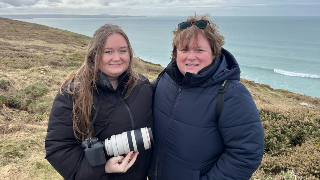 Two women, a mother and daughter, stood on a coast path with the sea behind them. They are both wearing puffer coats and the woman on the left is holding a digital camera with a long lens attached