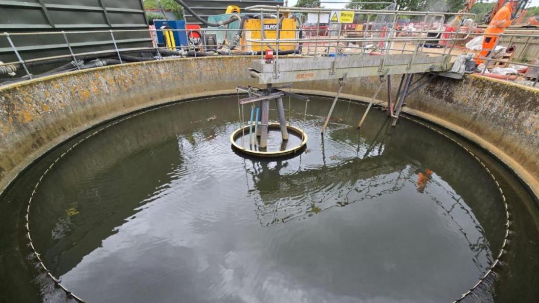 Large round tank of water with steep sides and workers and machinery above.
