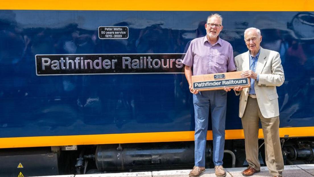Pathfinder Railtours boss Barry Cordell with founder Peter Watts. They are stood in front of a train which has the name pathfinder tours on its side. They are holding a miniature of the name in their hands.
