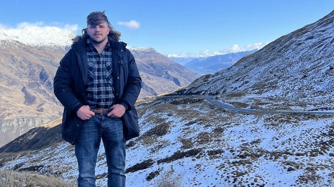 Percy who has a brown mullet looks at the camera, he wears a blue lumberjack shirt with a navy coat and he stands on a mountainous landscape in New Zealand.