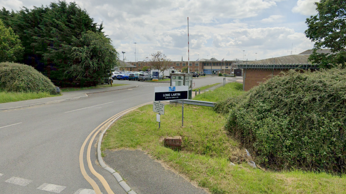 A road junction into Long Lartin Prison with a black and white Long Lartin sign in the grass near a bush. A car park can be seen in the background along with multiple brick buildings.