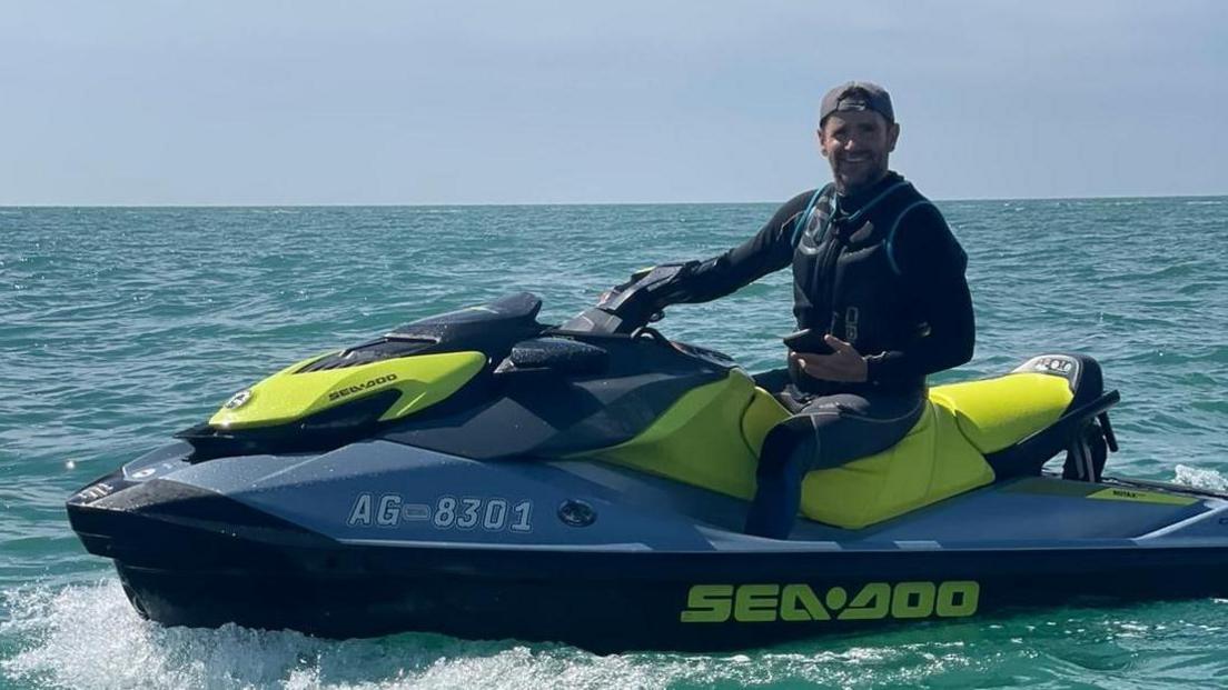 Mr Hopson is sat on a blue and yellow water scooter on a body of water. He is smiling and wearing a backwards cap and lifejacket. 