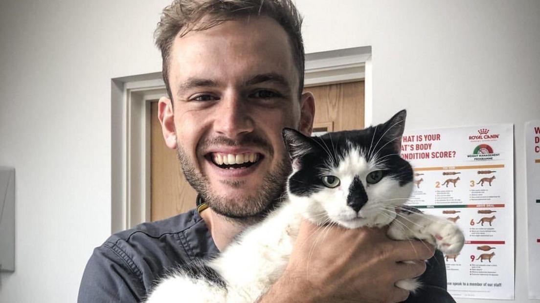 Vet Rory Cowlam who is smiling while holding a black and white cat