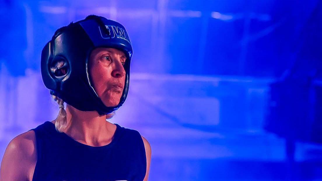 Teresa wearing a blue sleeveless boxing jersey and a blue helmet standing inside the boxing ring with blue lights.