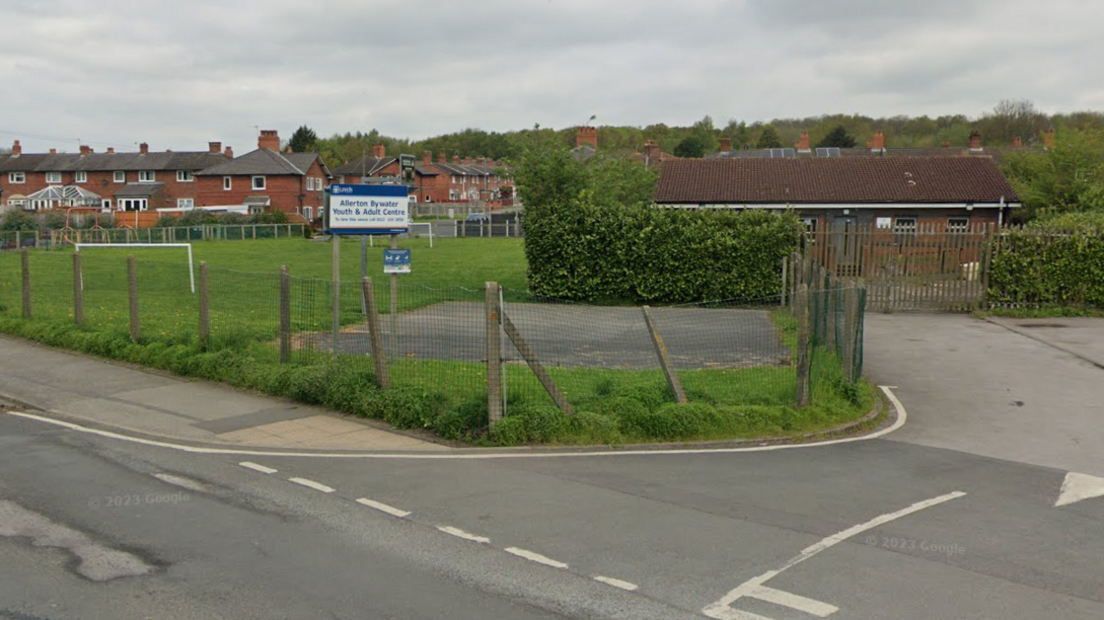 A car park and playing fields surrounded by a mesh fence with a sign reading "Allerton Bywater Youth and Adult Centre", which stands in the background.