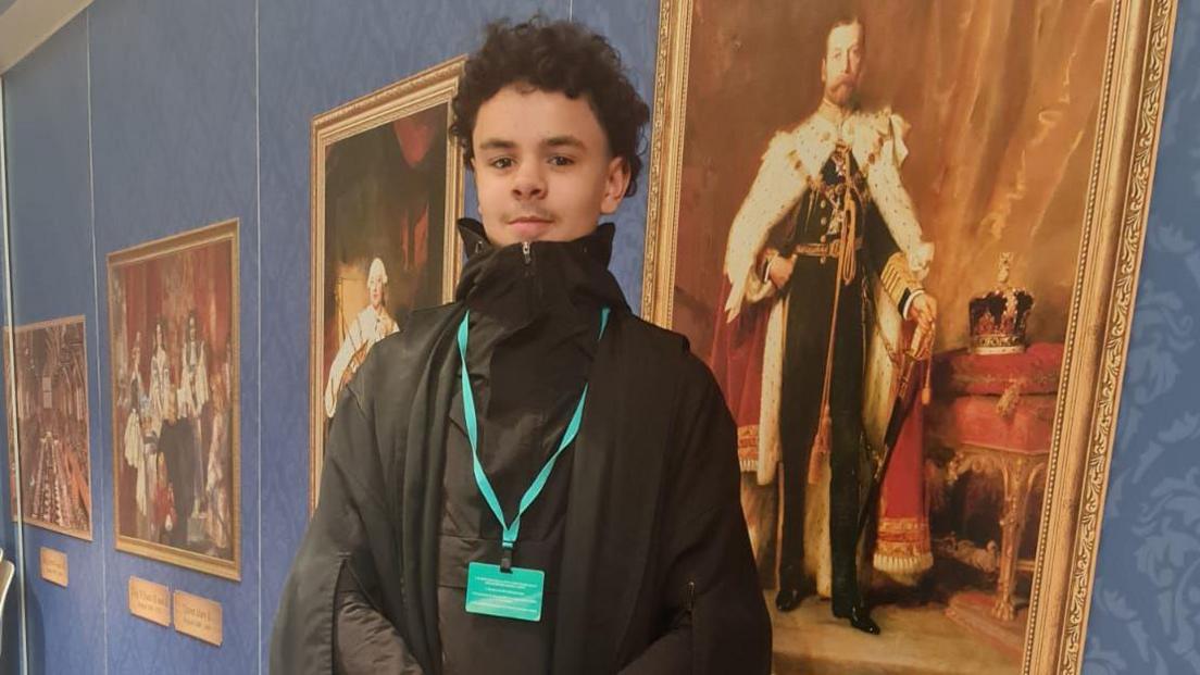 A picture of Darrian. He is wearing a black coat zipped all the way up and a black gown over it, with a green lanyard. He has curly dark hair and a thin moustache. He is standing in a display hall with painted portraits of kings and important figures behind him, on what looks like a school trip. 