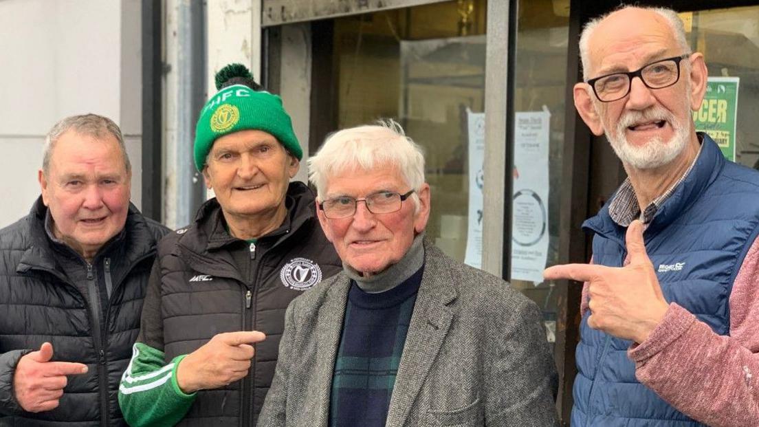 Four men pictured outside the trophy shop. 
Herbie Wade (Left) is in a black jacket, Gerry Doc has a green hat and black and green jacket, John 'Jobby' Crossan in a grey suit and jumper and Harry Nelis is pictured in a blue puffer jacket and is wearing glasses.