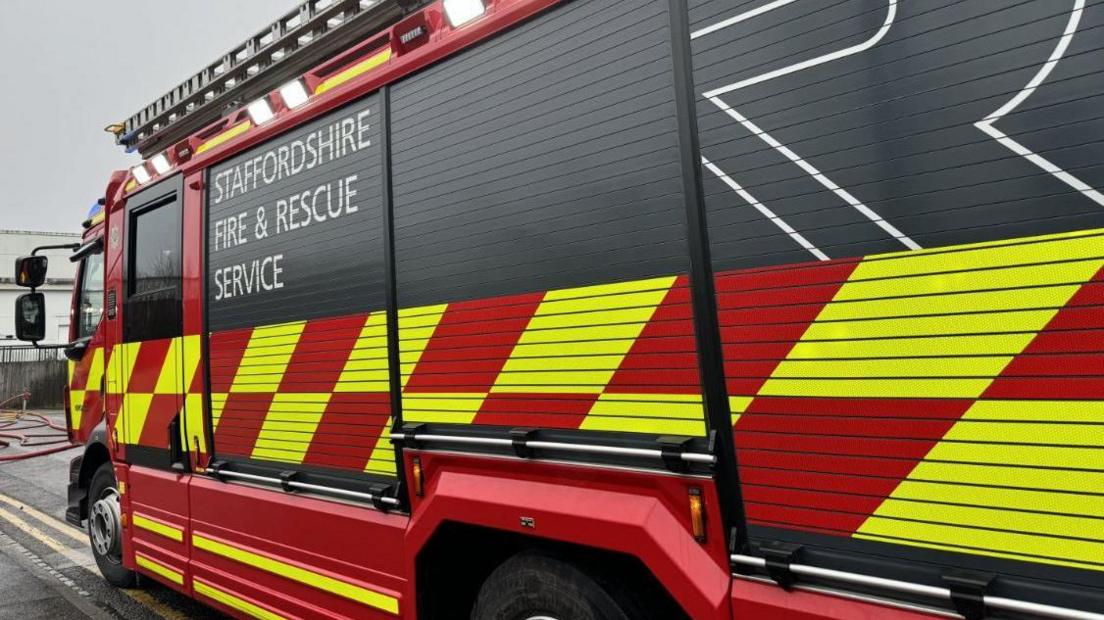 A side view of a Staffordshire Fire & Rescue Service appliance, with bright yellow and red chequered insignia and a ladder laid flat on the roof, parked outside.