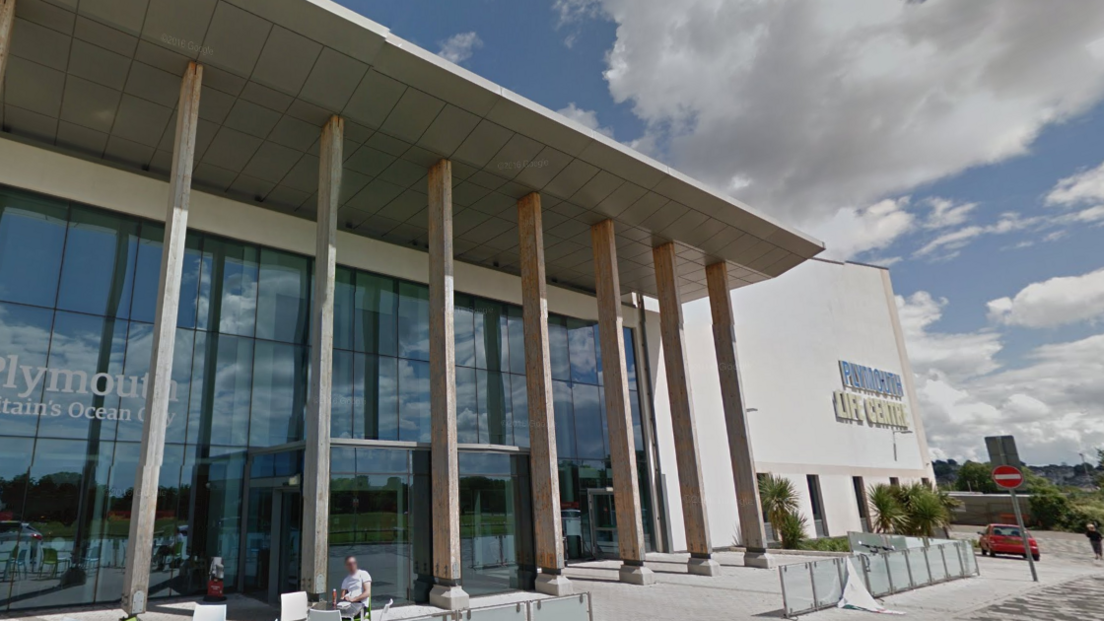 The outside of Plymouth Life Centre. It is a large glass fronted building with seven pillars propping up an extended solid awning. There is cloudy sky in the background of the image.