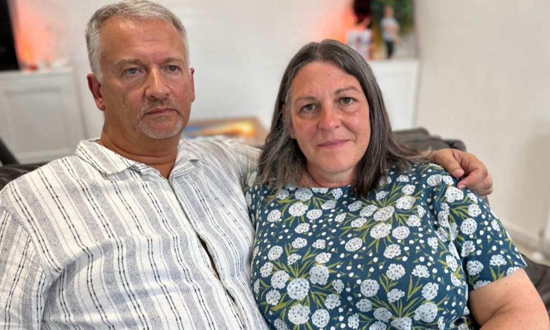 Middle aged man and woman sitting in a chair. The man is looking off the camera, the woman is facing it. The man has his arm around her shoulder.