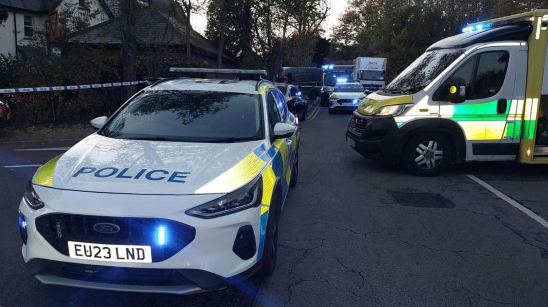 Two white police cars and an ambulance parked on a road with blue lights on. Blue and white police tape surrounds the vehicles at the scene. In the background vehicles can be seen queuing on what looks like a residential street, with trees both sides of the road and a house to the left.