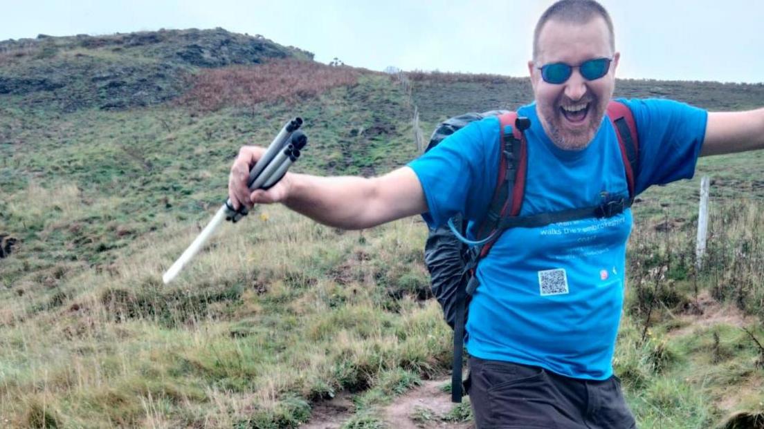 Chris Owens stands on a grassy hill with his arms raised to his side, with a folded walking stick in his right hand. He is smiling widely, wearing sunglasses, a bright blue t-shirt, black shorts and a backpack.