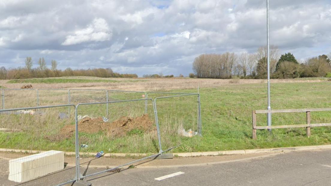 A road is at the bottom of the image which has a silver fence on the left blocking off parts of the road, and a wooden fence to the right. Behind them is a field which has trees on the other side in the background.