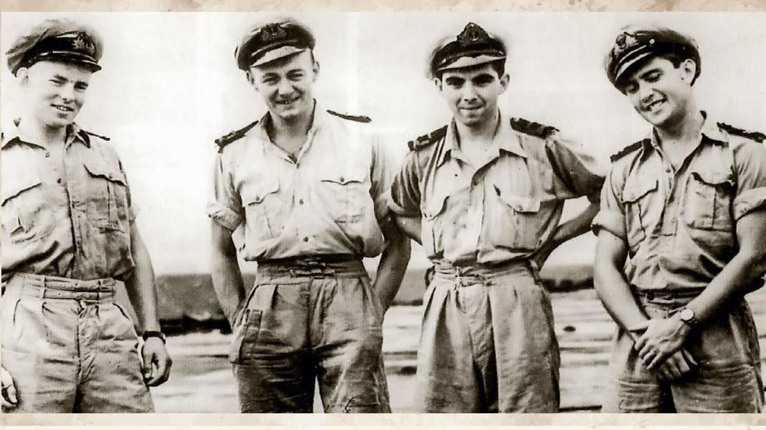 A black and white photograph of four young men in army uniform. They have their sleeves rolled up and are wearing military caps. They are standing around looking relaxed and are smiling.
