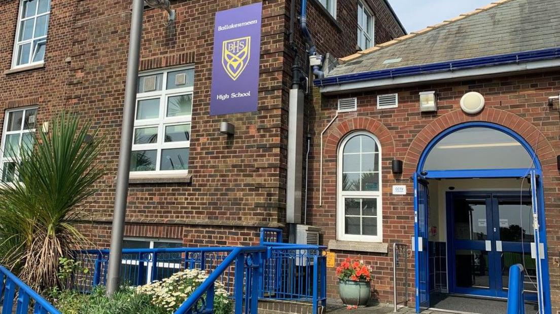 The entrance to Ballakermeen High School, which is a red-and-brown brick building with a blue-framed glass door and blue railings leading up to it.