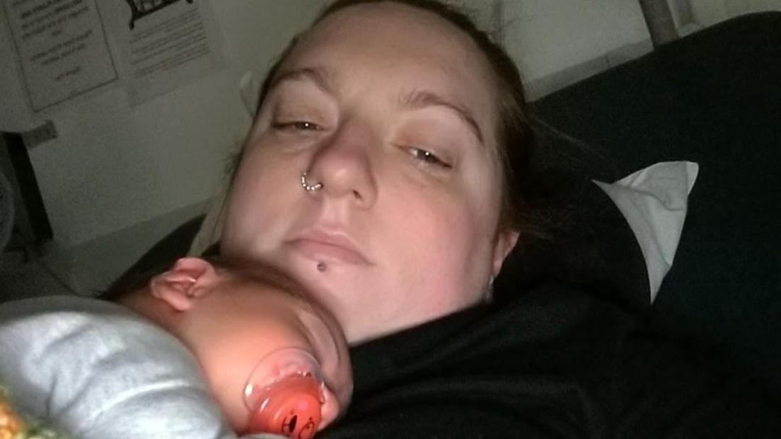 Debbie McCrudden lying on a dark coloured bed holding her baby who has a dummy in his mouth.