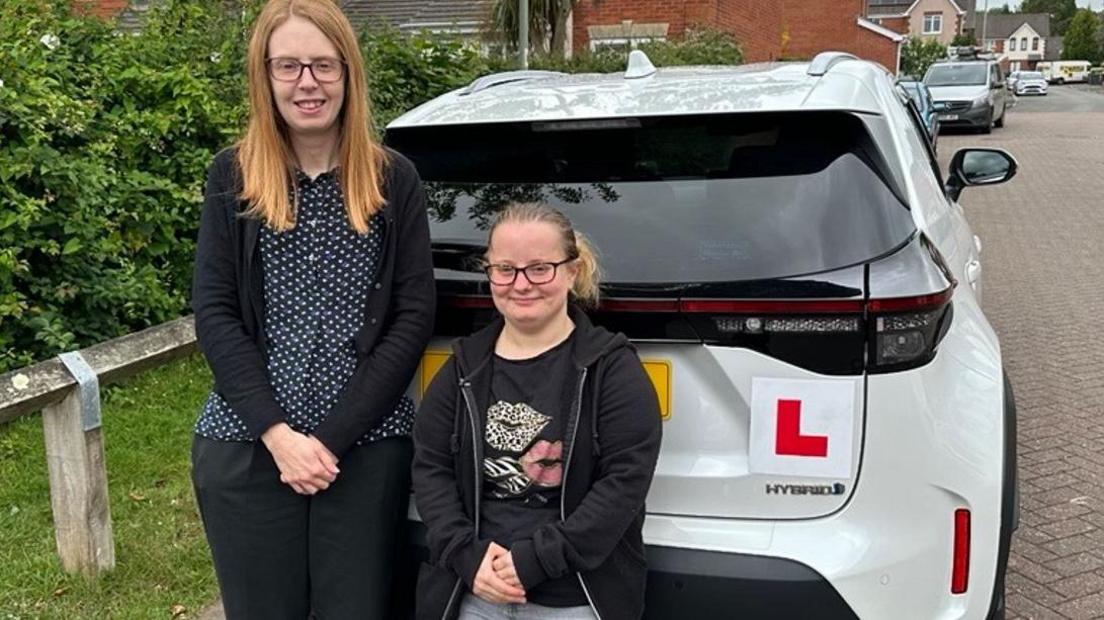 Chelsea Moon, who has a bone condition that stunts growth, waited a year for lessons with instructor Emma Hallett (left). Chelsea is just over four feet tall and is wearing a black hoodie, grey jeans and glasses. Emma, also in glasses, is wearing a black cardigan trousers and polka dotted shirt. They are standing behind a white car with learner plates.