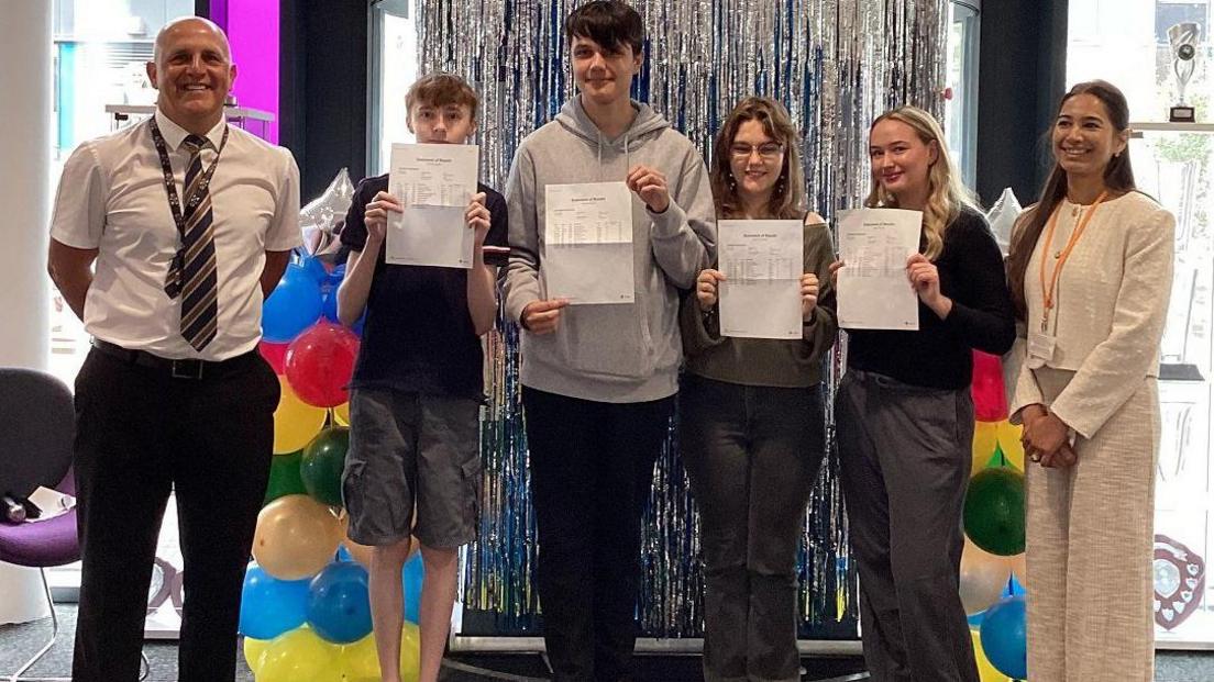 Students Charlie Williams, Sam Holicka, Joanna Marciniak, Sienna Keenan, with Jon Young, Executive Principal of The Deanery Church of England Academy, and Cabinet Member for Education, Councillor Adorabelle Shaikh. They stand in a line, the students holding up their results. All are smiling at the camera. There are balloons behind them.