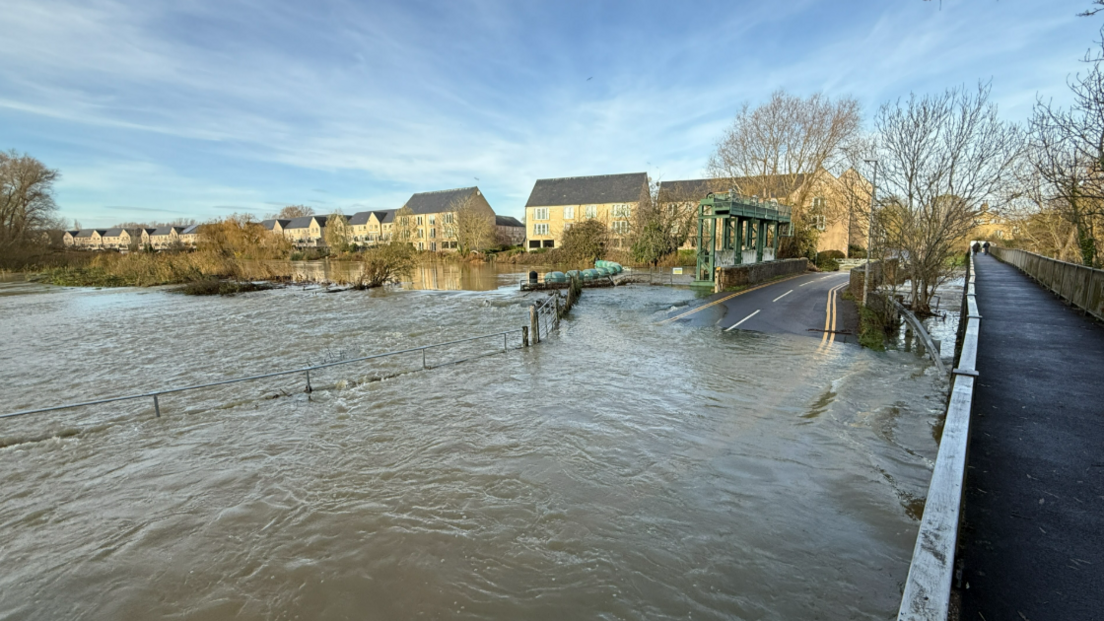 The River Great Ouse has burst its banks and flooded the road. Only the top of the bridge can be seen. Some double yellow lines can also be noticed in the murky brown water. 