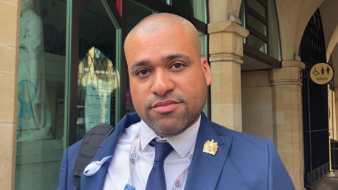 James Hill, who has close-cropped, receding grey hair and is wearing a blue suit with a blue tie and white shirt, stands outside the council offices. 