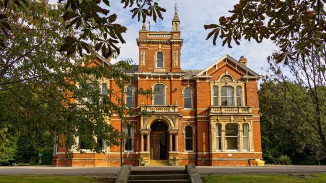 Weelsby Hall with trees and a lawn with steps leading up to where the building is. It has an orange exterior with beige bay windows and is three floors high.