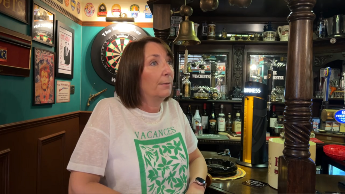 Gina Marchant with short dark hair and a green and white t-shirt stands leaning on the bar of the pub with the dartboard on the wall behind her