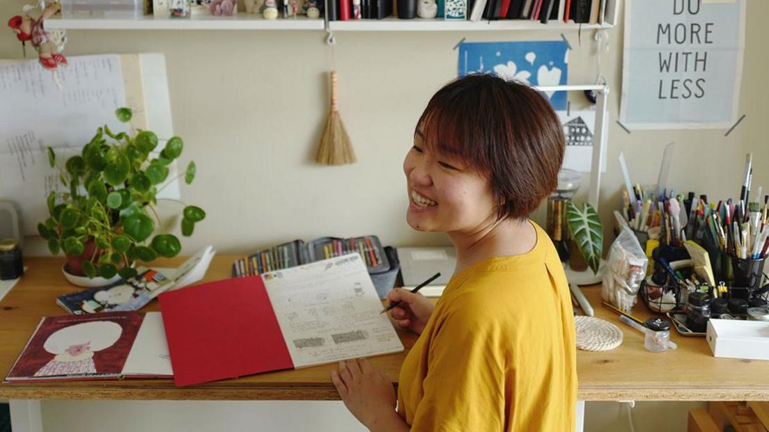 Woman wearing a yellow jumper is smiling with a pencil in her hand at a work desk. She has an illustration pad open. There are plants on the table and colouring pencils. A book shelf above the desk holds books and ornaments.