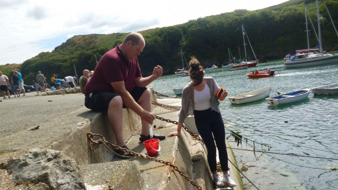 A photo of a young woman and a middle-aged man at a harbour. The man is sat down on some steps, wearing a maroon polo shirt and black shorts. The young woman has brown hair pulled into a bun and is wearing a white shirt, grey cardigan, and black jeans. Her hair obscures her face. Several boats are seen in the harbour.