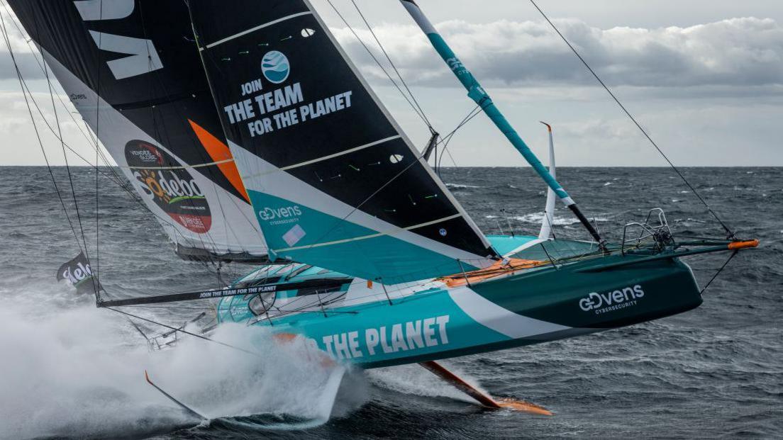 A racing yacht with a white and blue hull and black and white sails going fast with spray behind.