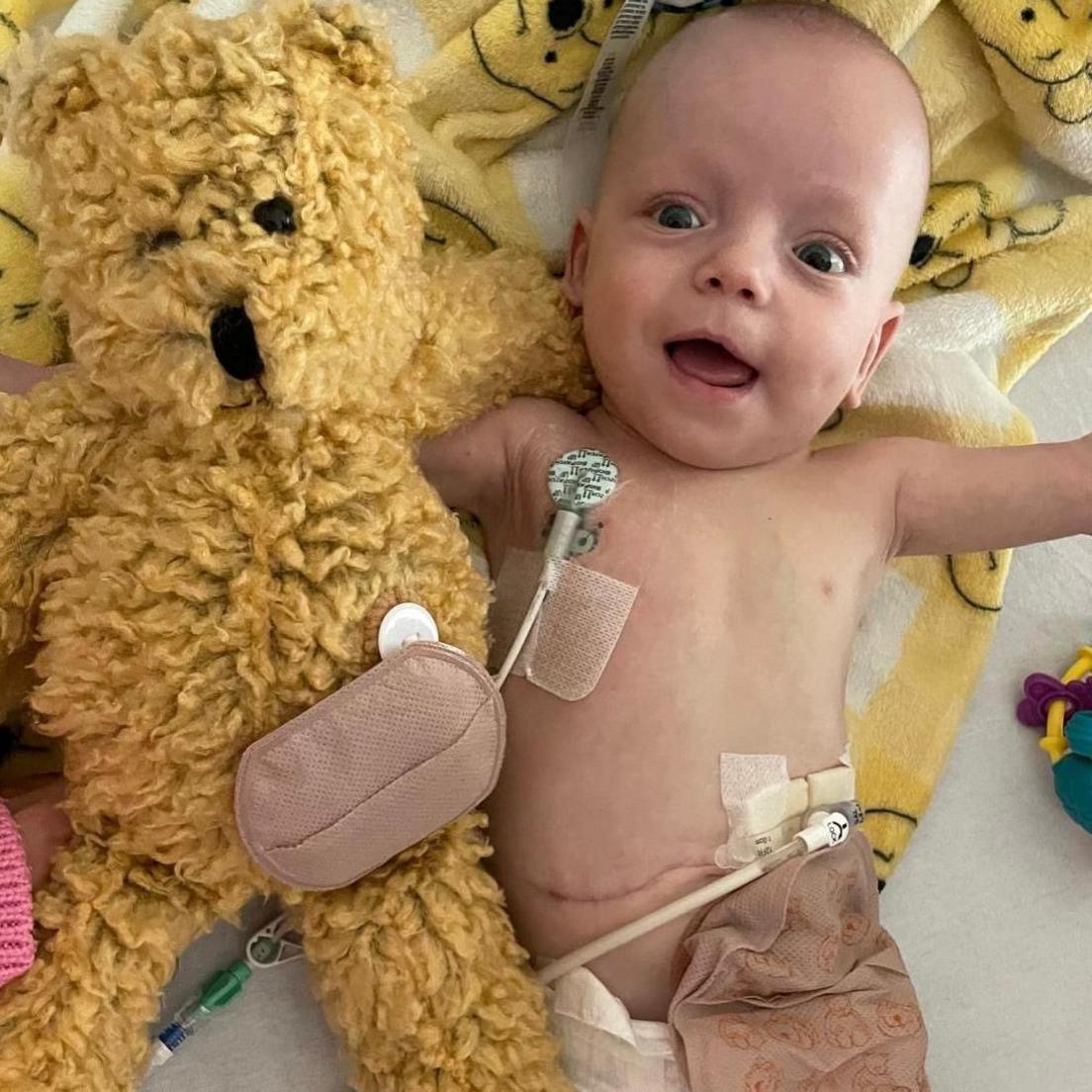 Baby Zachary in a hospital cot with monitors attached and lying beside a big yellow teddy bear