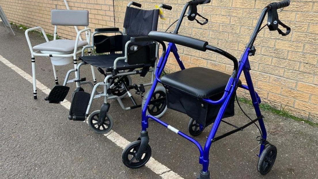 Mobility aids are sitting outside against a brick wall. There is a grey commode, a dark blue wheelchair and a black and blue walking aid with three wheels.