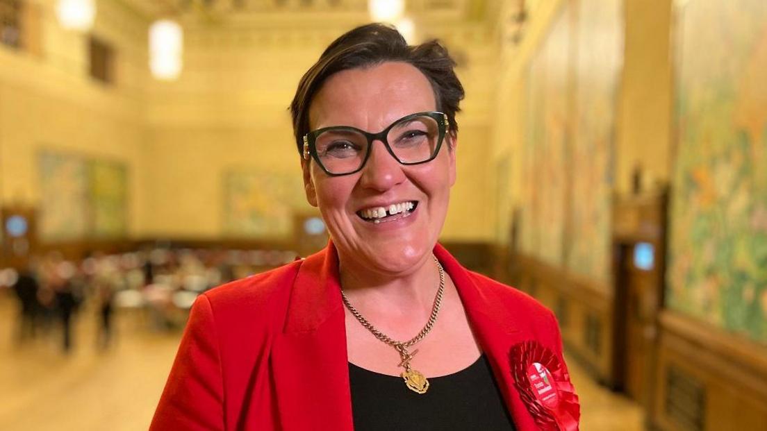 Tonia Antoniazzi is smiling to the camera while wearing a red suit jacket, black top and red Labour rosette. She has short dark hair parted to the side and large black-framed glasses 