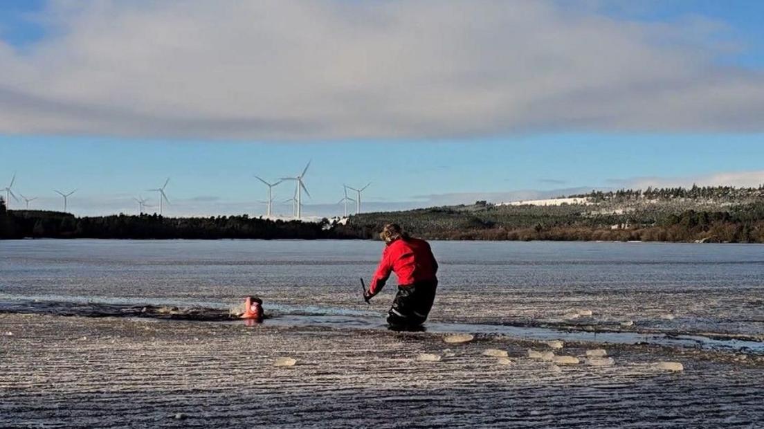 Frankie, with a swimming cap and goggles, swims through a channel in the icy Sweethope Lough. Coach Fenwick Ridely is standing in the water, knee deep, in a red coat and fluffy hat and monitoring her progress.