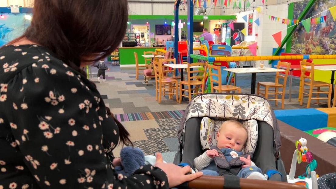 A woman in a black, flower-patterned dress, looks at a baby in a pushchair.