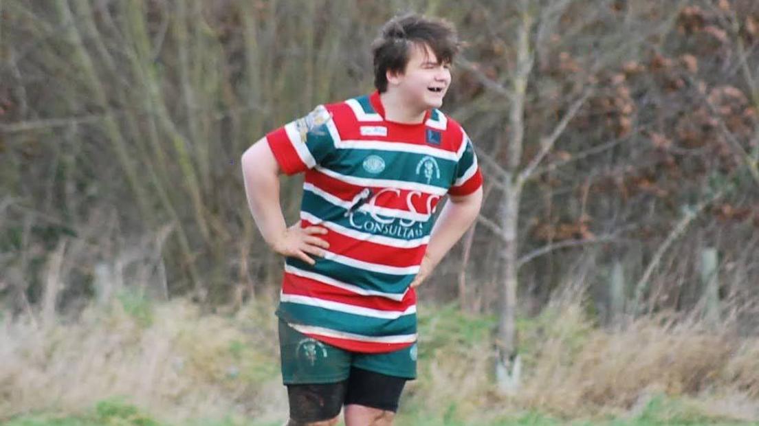 A teenager with brown windswept hair wearing a stripy green, red and white shirt with his hand on his hips and looking into the distance. He is also wearing green and black shorts and a woodland can be seen behind him.