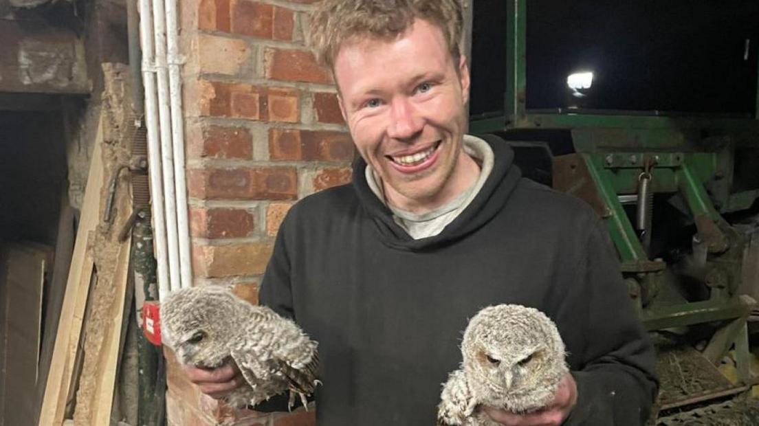 Carruthers, with short blond hair and wearing a black hoody, smiles as he holds two young grey owls, one in each hand. Behind him is a wall and agricultural equipment