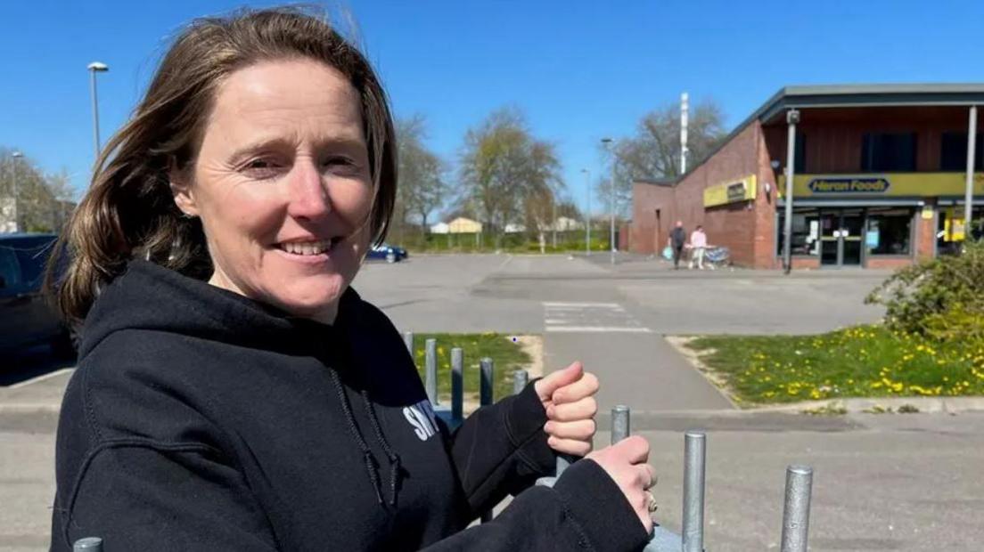 Jo Lorenz, wearing a black St Michael's Youth Project hoodie, smiles for the camera. In the background is a single-storey shopping parade