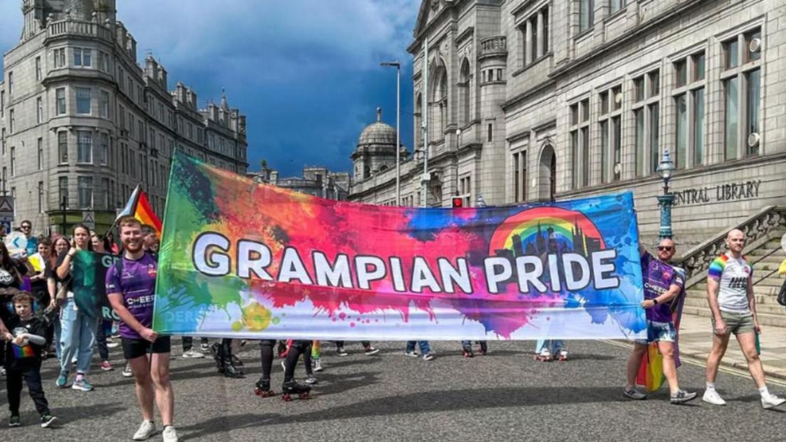 People attending Pride with a colourful banner which says "Grampian Pride". It has a rainbow and outline of Aberdeen's skyline.