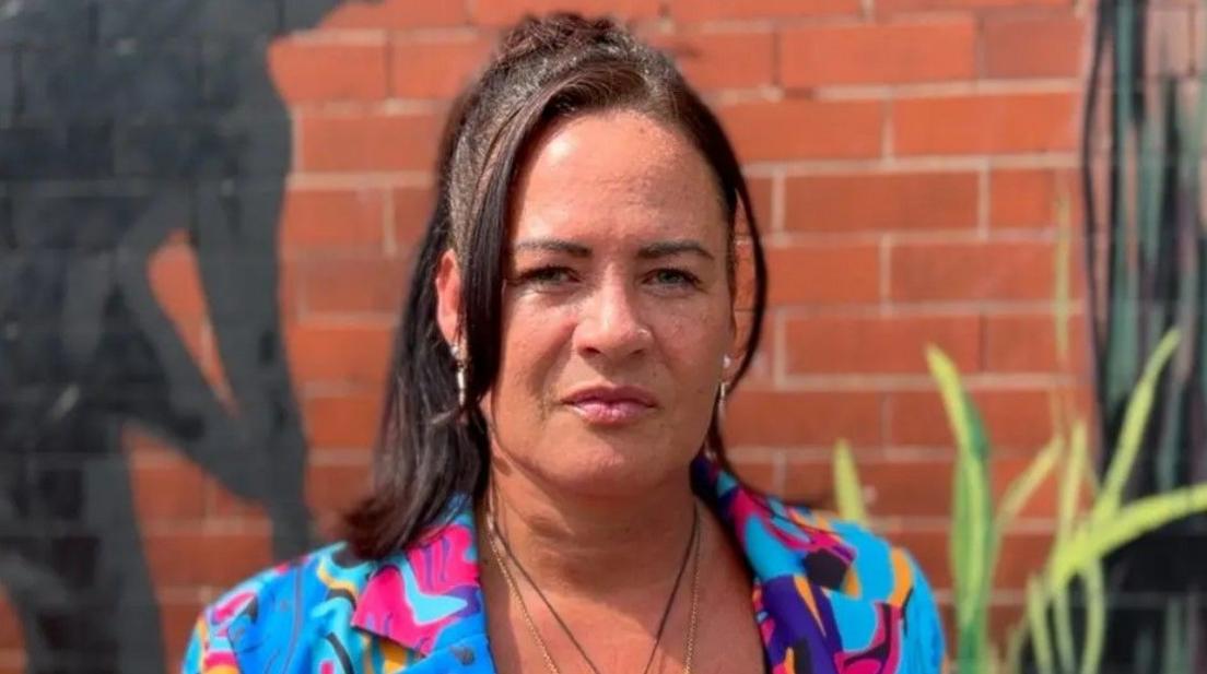Kelly Brown with a brightly coloured patterned jacket stands in front of a mural on a brick wall.
