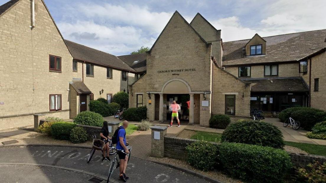 Outside the Witney Four Pillars Hotel on a cloudy day. A couple of people are seen on bicycles in front of the building. Another man is standing at the entrance.