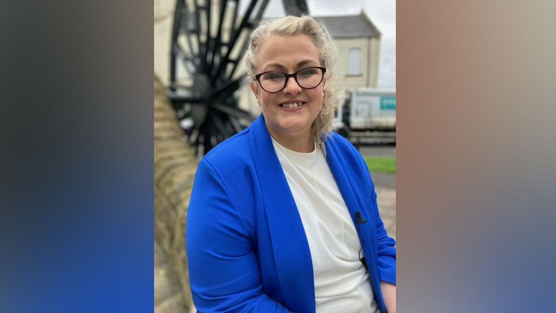 Ms Howey, dressed in a blue blazer, is sitting and smiling at the camera. She's wearing a blue blazer and a white top. She has blonde hair, which is in a ponytail and wears glasses.