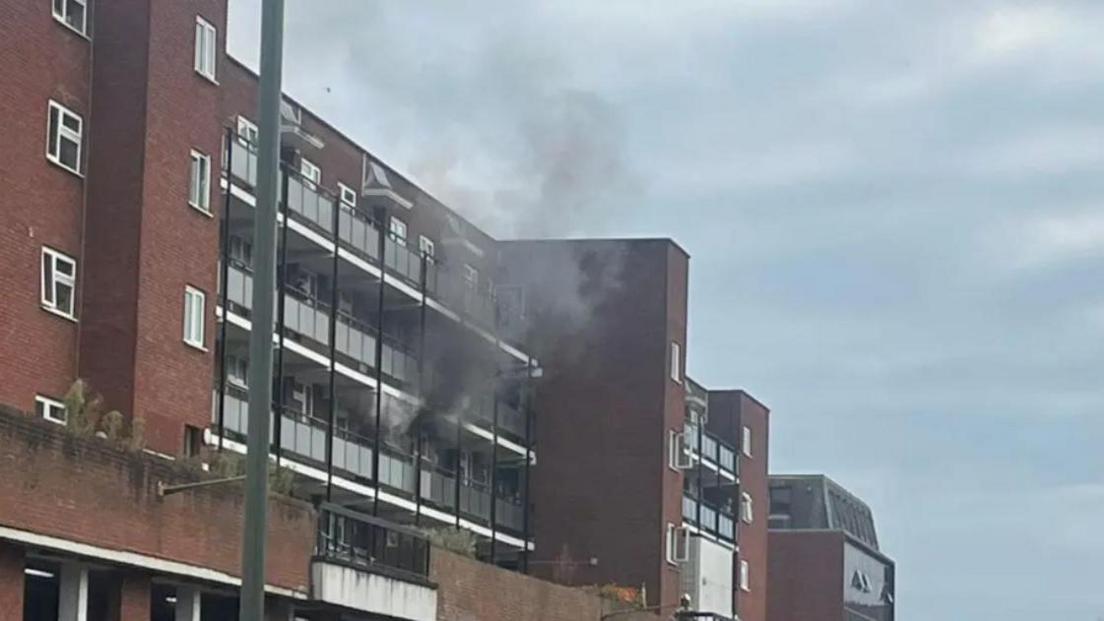 Exterior of a four-storey block of flats with black smoke pouring out of one of the windows over balconies on second floor