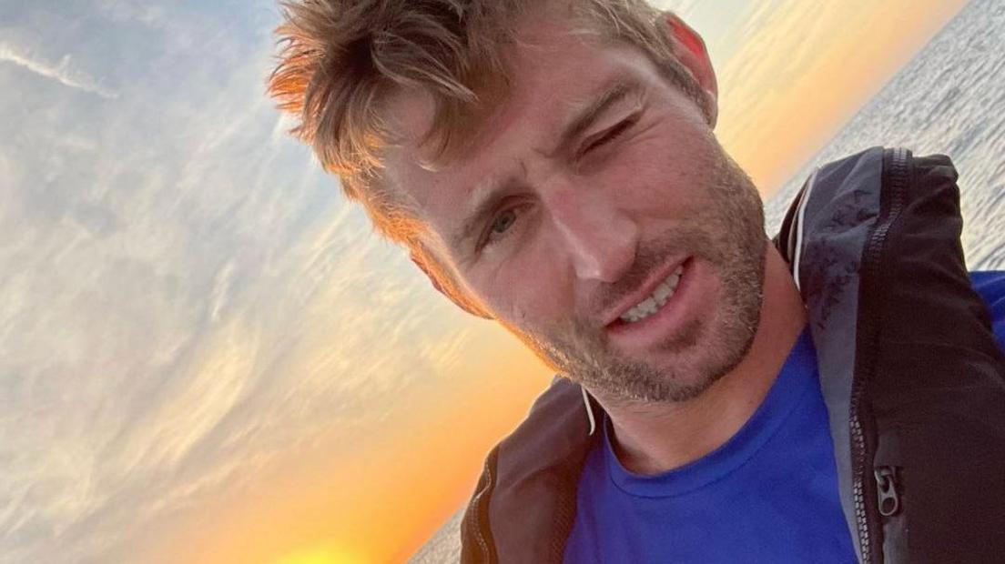 Tom Waddington taking a selfie with his left eye shut, wearing a blue T-shirt and black life jacket with a sunset and the ocean behind him