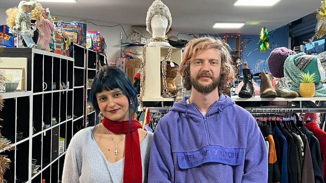 Ashira Denab, who has black hair with a bluish tinge and is wearing a light blue sweater and dark red scarf standing next to Rick Hartley who has blonde hair, a dark beard and a blue hooded fleece. They are in front of a clothing rail inside a charity shop which has been decked out with Christmas decorations. 