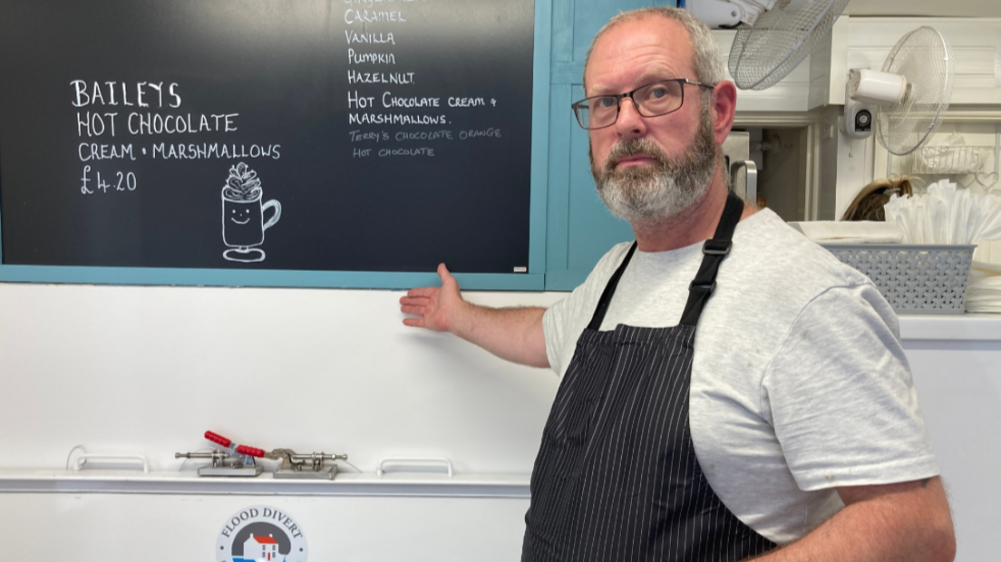 A man wearing a black striped apron points to just under a menu board on a wall