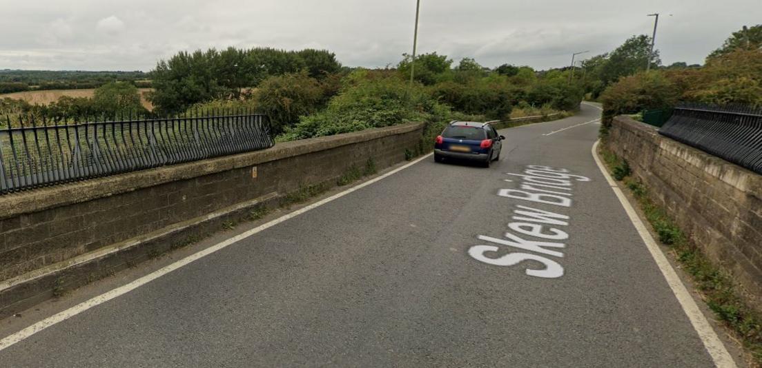 An image of a country road. A blue car is visible. Railings are visible to the right and left where the road crosses a railway line, but the line itself is not in the picture. 