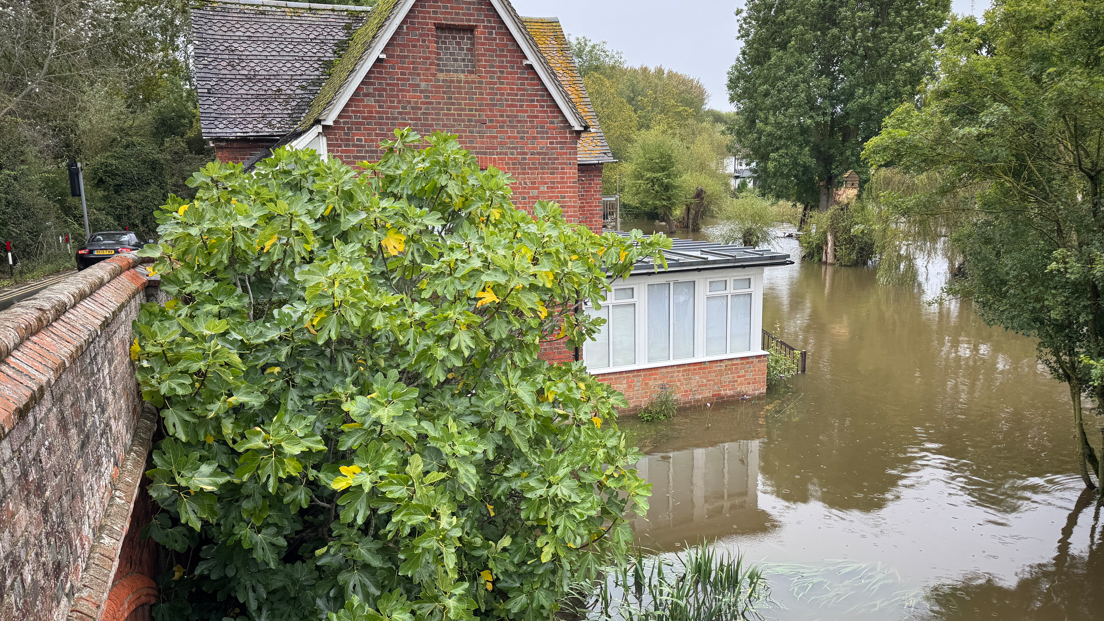 A home surronded by flood water. The water is brown and murky. Trees can be seen around the home.