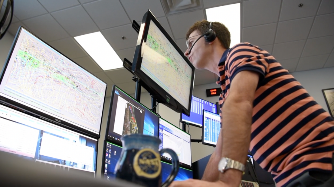 photo of a man standing at a desk, talking into a microphone while looking at numerous computer screens